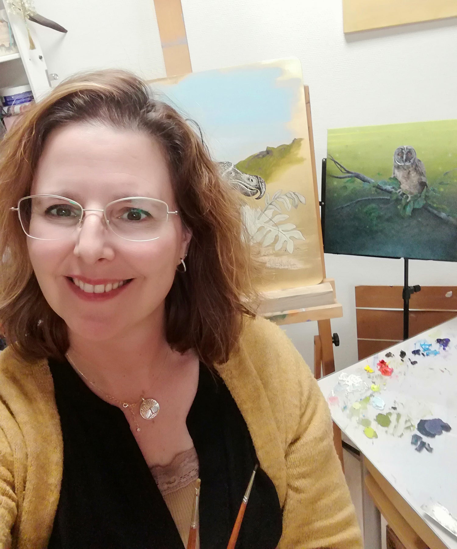 Portrait photo of Cindy Sangen in her home Art studio. On the background two artworks, paintings Cindy Sangen is working on. Palet with oilpaint and brushes are in view.